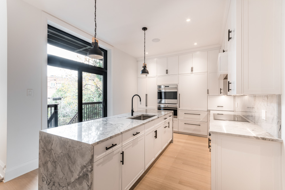 Photo of a large arts and crafts u-shaped kitchen pantry in Montreal with shaker cabinets, white cabinets, granite benchtops, grey splashback, stainless steel appliances, light hardwood floors, with island and grey benchtop.