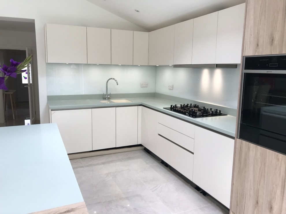 Design ideas for a mid-sized modern l-shaped eat-in kitchen in Kent with an undermount sink, flat-panel cabinets, white cabinets, solid surface benchtops, white splashback, glass sheet splashback, black appliances, porcelain floors, with island, grey floor and green benchtop.