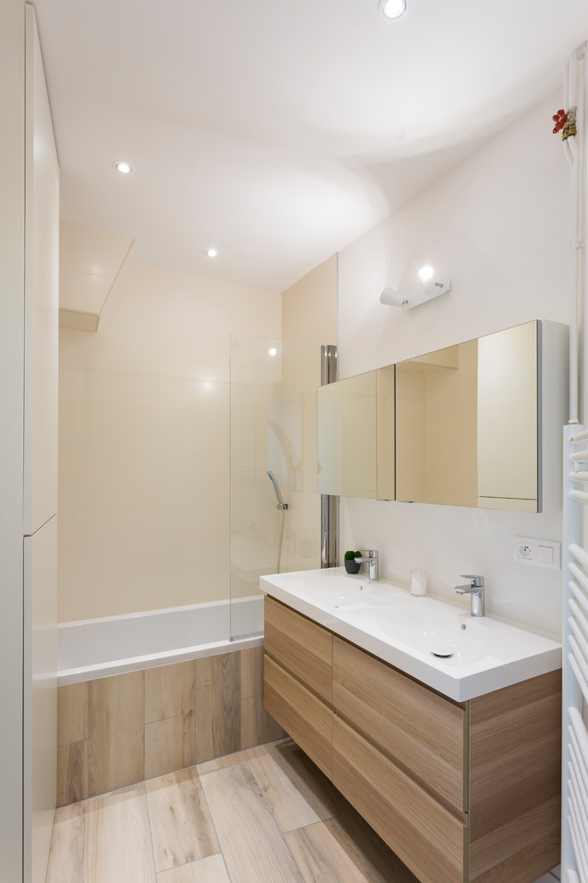 Small contemporary master bathroom in Paris with white walls, light hardwood floors, beige floor, flat-panel cabinets, light wood cabinets, a drop-in tub, a shower/bathtub combo, a two-piece toilet, beige tile, porcelain tile, a console sink, solid surface benchtops, a hinged shower door and white benchtops.