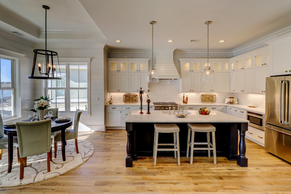 This is an example of a large country l-shaped open plan kitchen in Atlanta with an undermount sink, shaker cabinets, white cabinets, quartz benchtops, white splashback, subway tile splashback, stainless steel appliances, light hardwood floors, with island, brown floor and white benchtop.