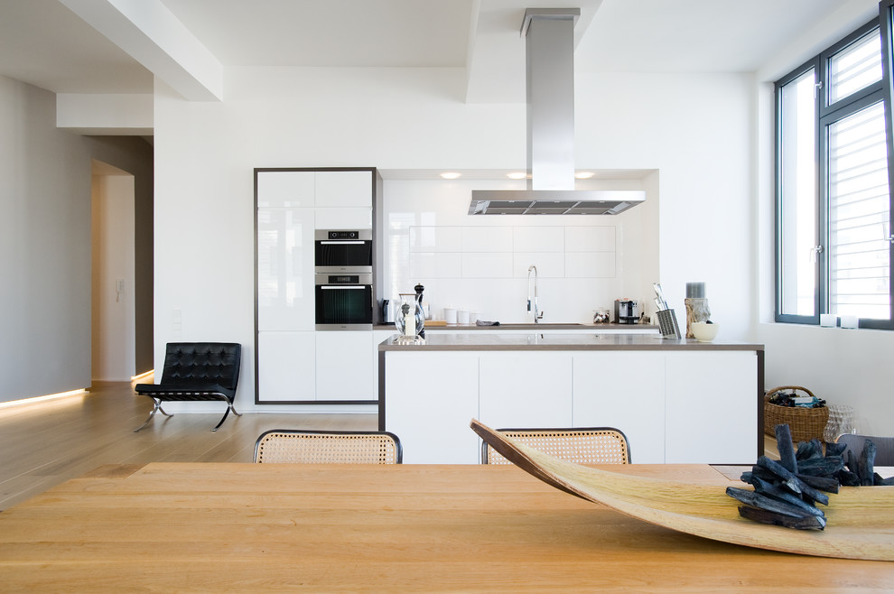 Photo of a mid-sized contemporary single-wall eat-in kitchen in Hamburg with flat-panel cabinets, white cabinets, white splashback, panelled appliances, light hardwood floors, with island and stainless steel benchtops.