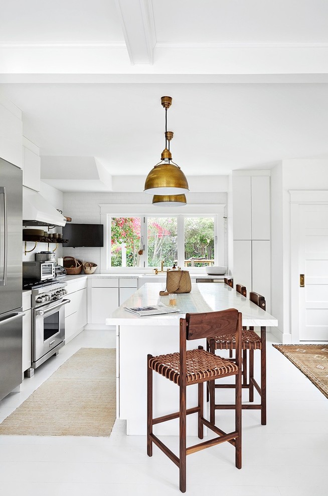 Beach style l-shaped kitchen in Los Angeles with a farmhouse sink, flat-panel cabinets, white cabinets, white splashback, stainless steel appliances, with island, white floor and white benchtop.