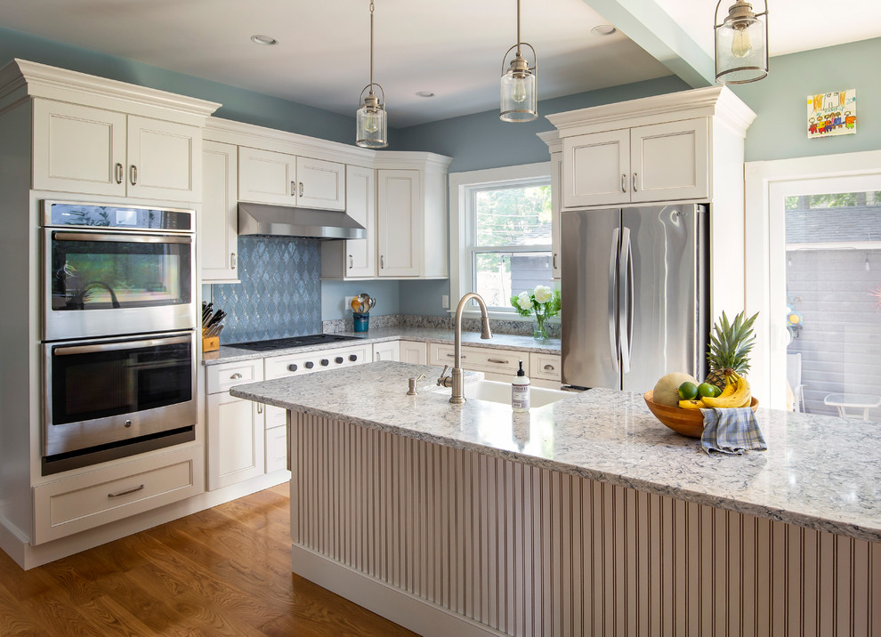 Photo of a mid-sized traditional l-shaped kitchen in Boston with a farmhouse sink, white cabinets, quartz benchtops, blue splashback, ceramic splashback, stainless steel appliances, medium hardwood floors, with island, white benchtop, recessed-panel cabinets and brown floor.