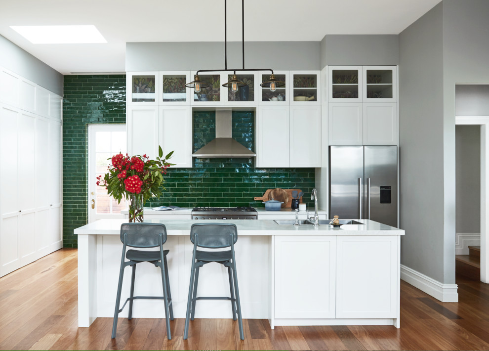 Inspiration for a transitional galley open plan kitchen in Sydney with a double-bowl sink, shaker cabinets, white cabinets, green splashback, subway tile splashback, stainless steel appliances, medium hardwood floors, with island, brown floor and white benchtop.