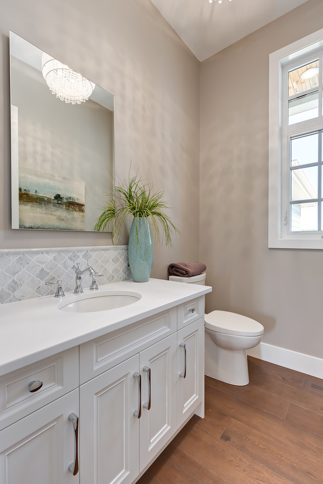 This is an example of a transitional powder room in Calgary with beaded inset cabinets, white cabinets, a two-piece toilet, gray tile, grey walls, medium hardwood floors, an undermount sink, brown floor and white benchtops.