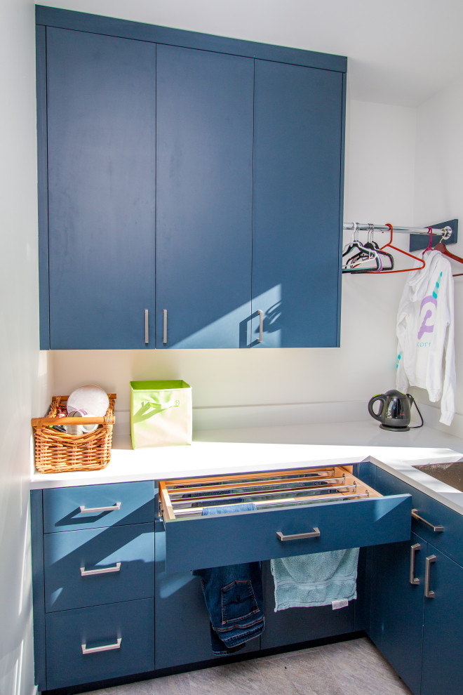 Photo of a mid-sized modern galley utility room in Minneapolis with an undermount sink, flat-panel cabinets, blue cabinets, quartz benchtops, engineered quartz splashback, white walls, a stacked washer and dryer and white benchtop.