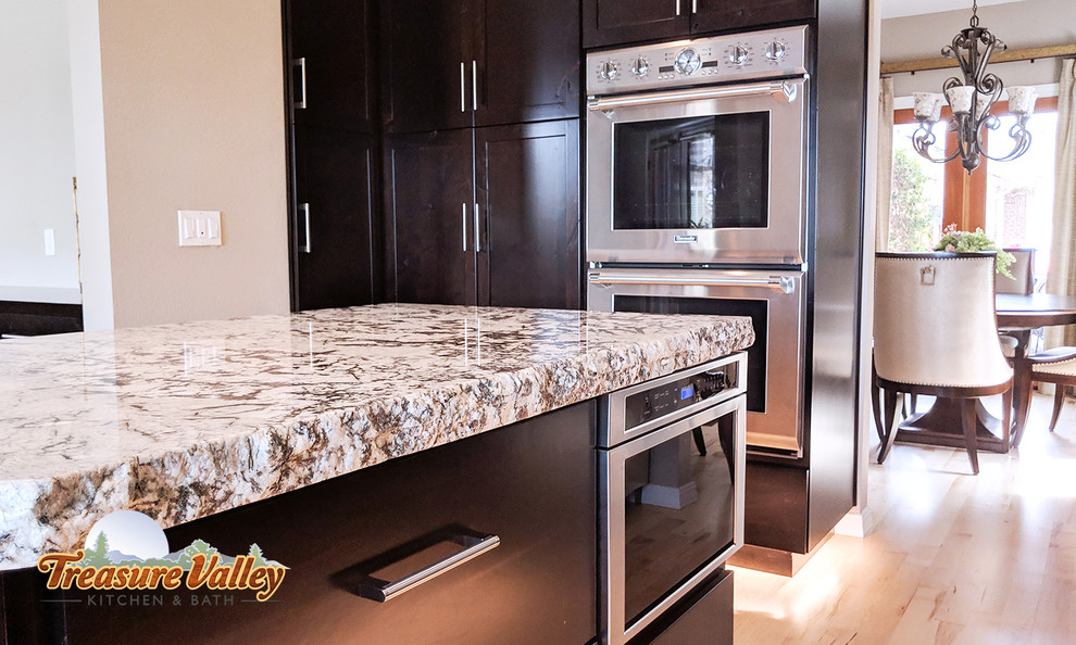 Mid-sized transitional l-shaped light wood floor open concept kitchen photo in Boise with a farmhouse sink, shaker cabinets, granite countertops, glass tile backsplash, stainless steel appliances and an island
