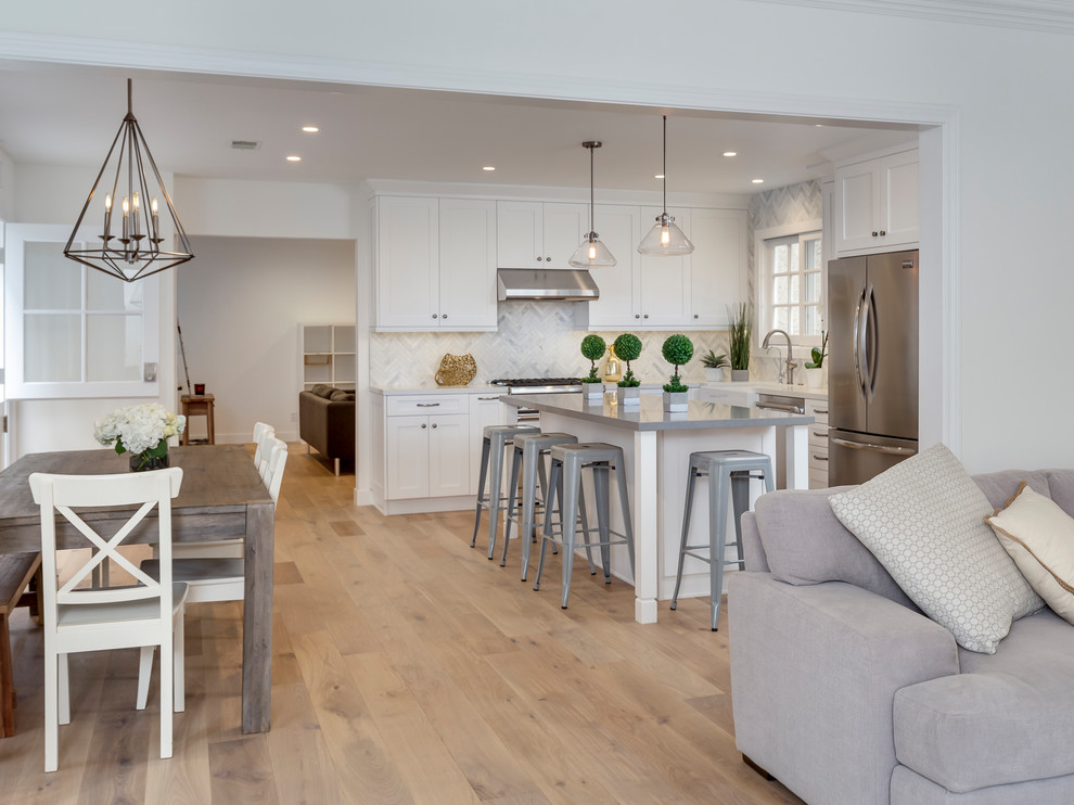 Inspiration for a large transitional l-shaped eat-in kitchen in Los Angeles with a farmhouse sink, recessed-panel cabinets, white cabinets, quartz benchtops, white splashback, stone tile splashback, stainless steel appliances, light hardwood floors and with island.