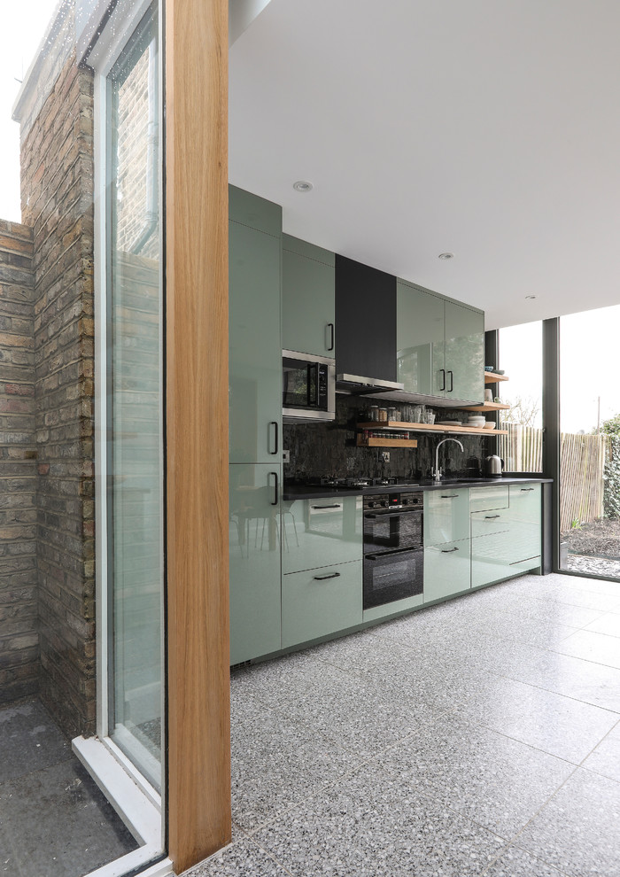 Design ideas for a mid-sized contemporary single-wall open plan kitchen in London with a drop-in sink, flat-panel cabinets, green cabinets, solid surface benchtops, black splashback, ceramic splashback, stainless steel appliances, terrazzo floors, grey floor and black benchtop.