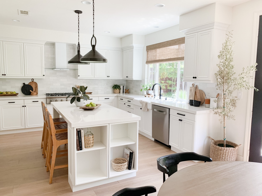 Example of a mid-sized transitional u-shaped light wood floor and beige floor enclosed kitchen design in San Diego with a farmhouse sink, raised-panel cabinets, white cabinets, quartz countertops, white backsplash, ceramic backsplash, stainless steel appliances, an island and white countertops