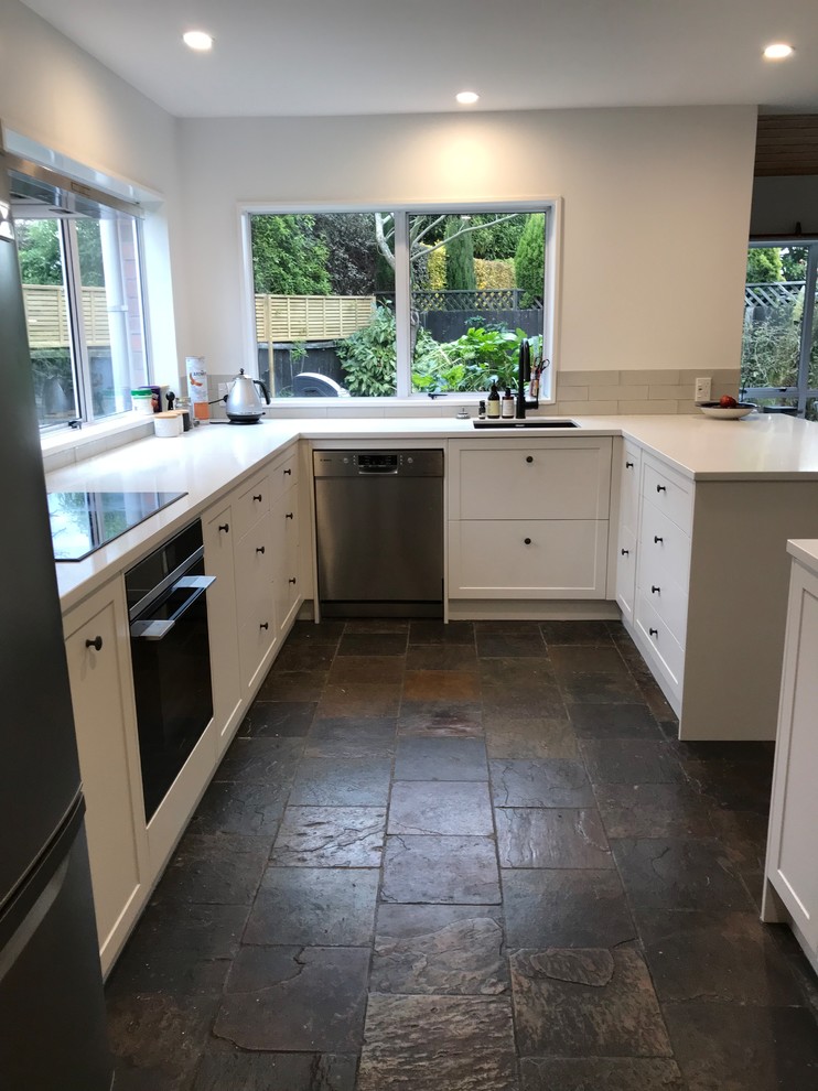 This is an example of a large contemporary u-shaped open plan kitchen in Christchurch with a single-bowl sink, recessed-panel cabinets, white cabinets, quartz benchtops, grey splashback, subway tile splashback, stainless steel appliances, slate floors, a peninsula, black floor and white benchtop.