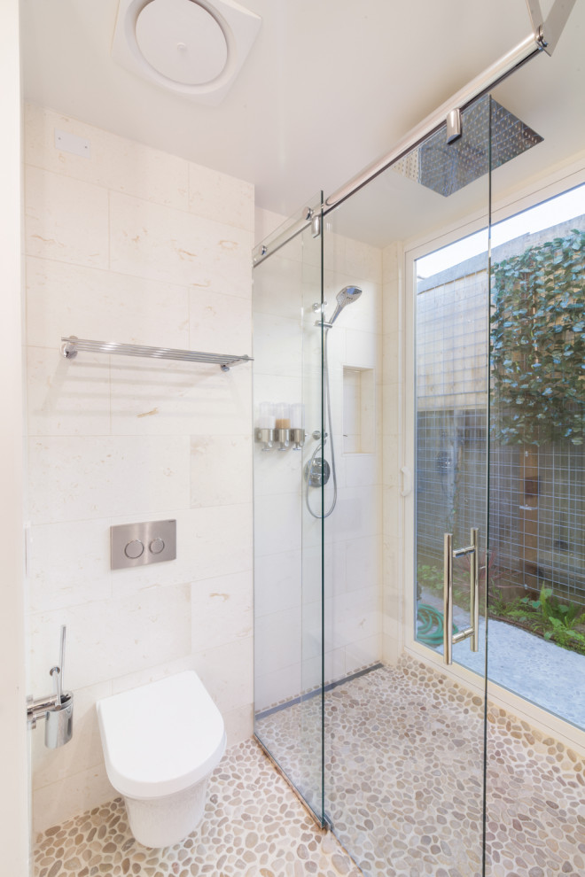 Photo of a small contemporary master bathroom in Los Angeles with flat-panel cabinets, brown cabinets, a freestanding tub, a curbless shower, a wall-mount toilet, beige tile, pebble tile, beige walls, terrazzo floors, a vessel sink, marble benchtops, beige floor, a sliding shower screen and beige benchtops.