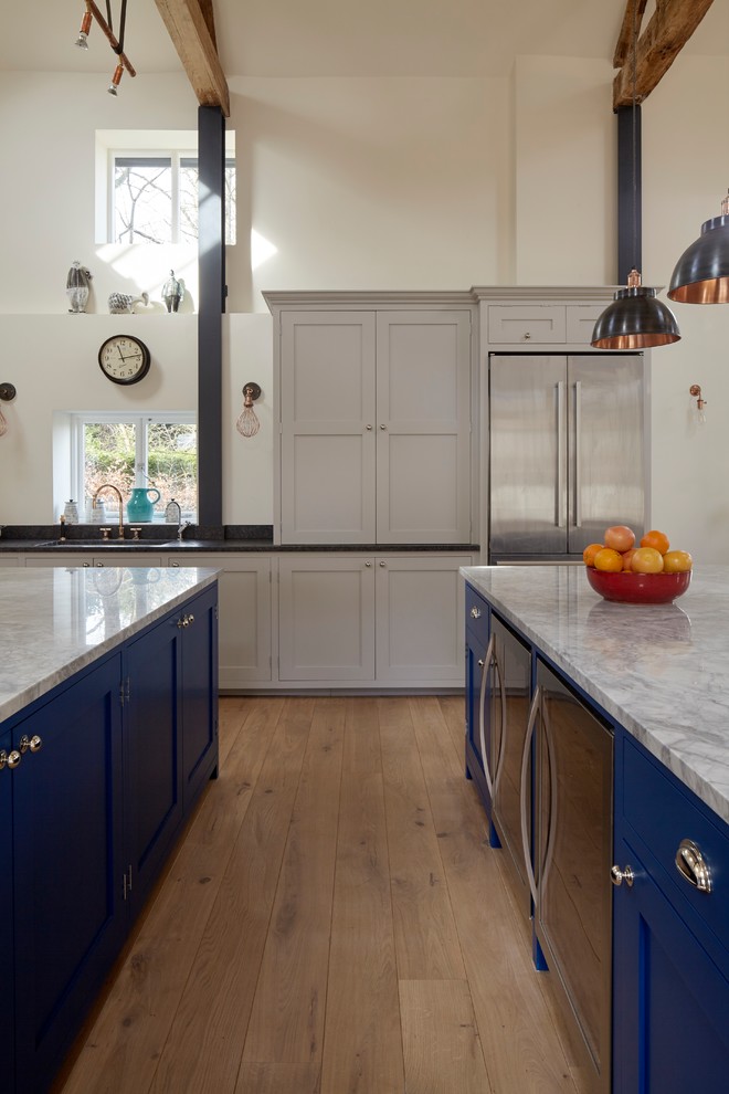 This is an example of a large traditional open plan kitchen in London with a drop-in sink, shaker cabinets, blue cabinets, marble benchtops, white splashback, subway tile splashback, stainless steel appliances, light hardwood floors, multiple islands and brown floor.