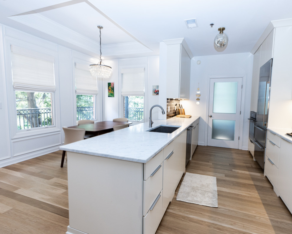 Photo of a large scandinavian galley eat-in kitchen in Atlanta with an undermount sink, flat-panel cabinets, beige cabinets, marble benchtops, black splashback, subway tile splashback, stainless steel appliances, light hardwood floors, with island, brown floor and white benchtop.