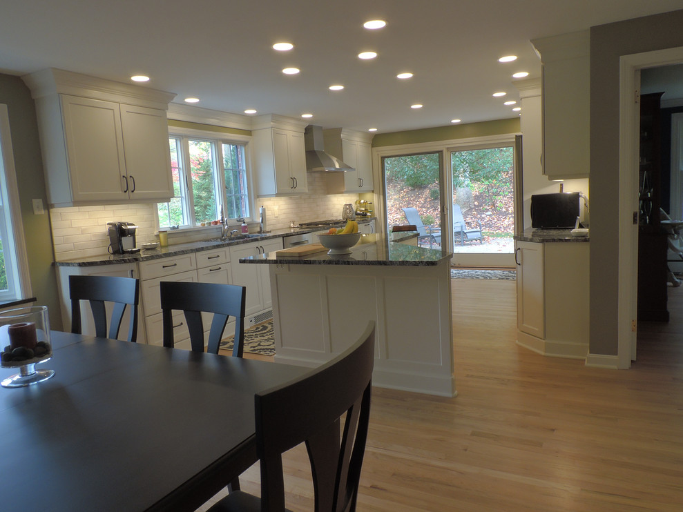 Example of a transitional light wood floor kitchen design with a single-bowl sink, shaker cabinets, white cabinets, granite countertops, white backsplash, ceramic backsplash, stainless steel appliances, an island and black countertops