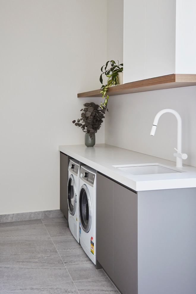Inspiration for a mid-sized contemporary galley dedicated laundry room in Melbourne with a drop-in sink, green cabinets, white splashback, porcelain splashback, white walls, ceramic floors, a side-by-side washer and dryer, grey floor and white benchtop.