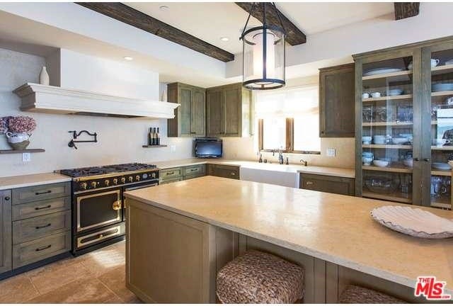 Photo of a large country u-shaped eat-in kitchen in Los Angeles with a farmhouse sink, glass-front cabinets, distressed cabinets, marble benchtops, black appliances, travertine floors and with island.