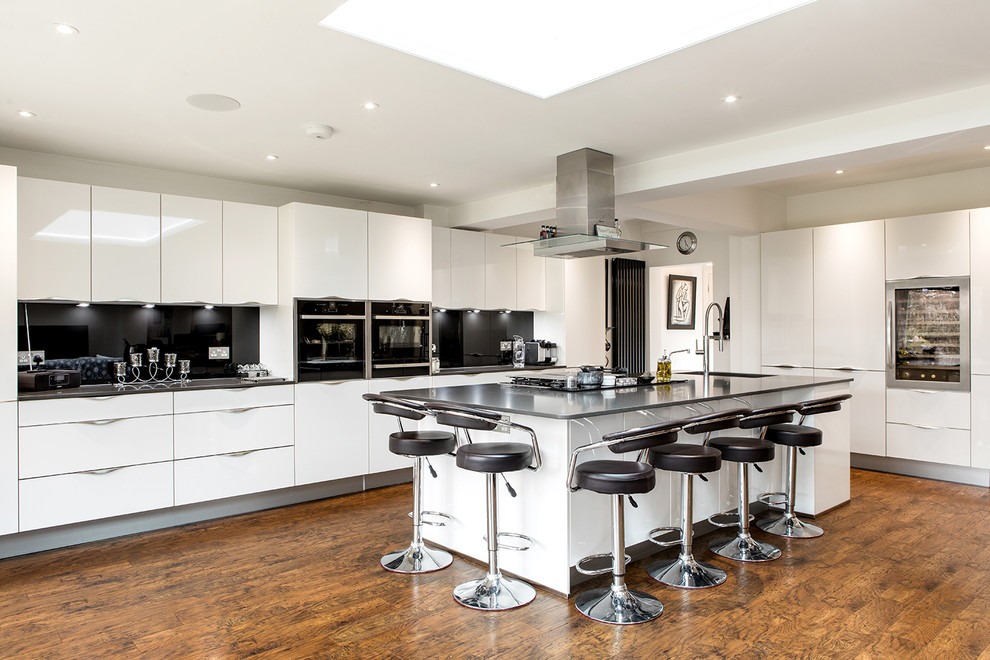Expansive contemporary l-shaped kitchen in London with an undermount sink, flat-panel cabinets, white cabinets, quartzite benchtops, black splashback, glass sheet splashback, panelled appliances, with island, black benchtop, medium hardwood floors and brown floor.