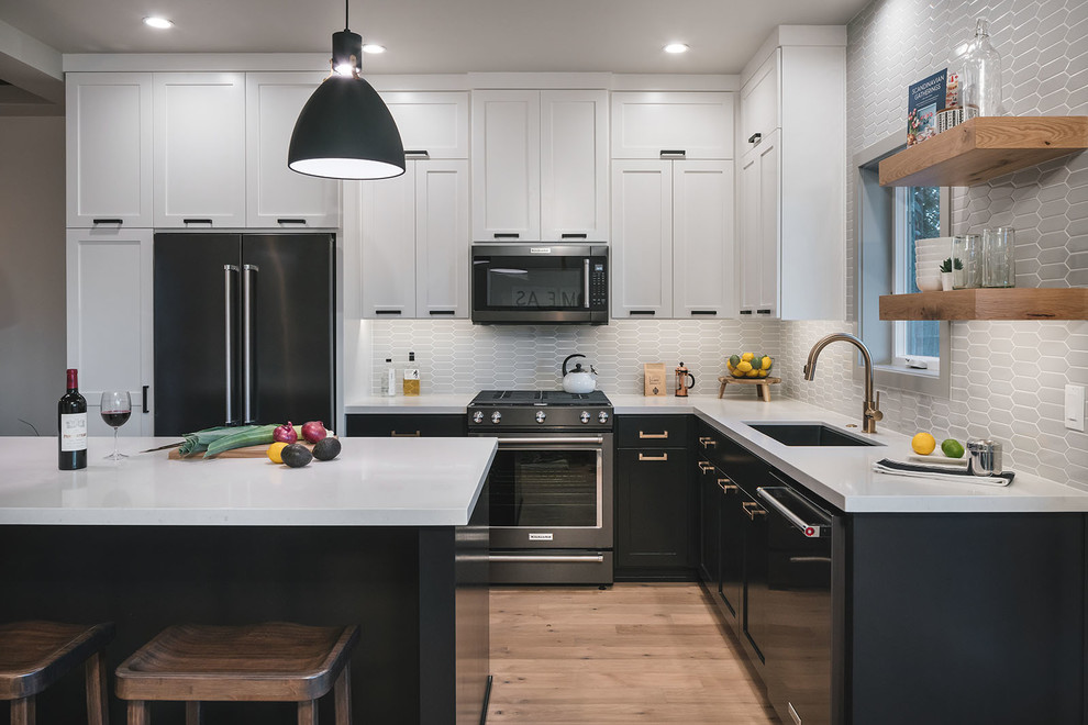 This is an example of a mid-sized transitional l-shaped open plan kitchen in Portland with an undermount sink, shaker cabinets, black cabinets, quartz benchtops, grey splashback, porcelain splashback, black appliances, light hardwood floors, with island, brown floor and white benchtop.