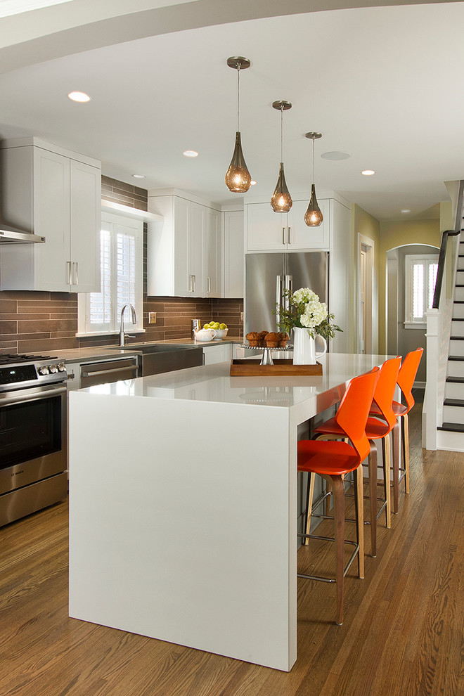 Inspiration for a mid-sized transitional l-shaped kitchen in Minneapolis with a farmhouse sink, shaker cabinets, white cabinets, quartz benchtops, stainless steel appliances, medium hardwood floors, with island and brown splashback.