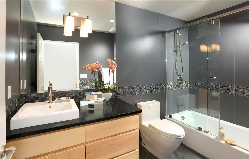 Mid-sized transitional 3/4 bathroom in Los Angeles with flat-panel cabinets, light wood cabinets, an alcove tub, a shower/bathtub combo, a one-piece toilet, gray tile, grey walls, porcelain floors, a wall-mount sink, soapstone benchtops, grey floor, a sliding shower screen, grey benchtops, a niche, a single vanity and a floating vanity.