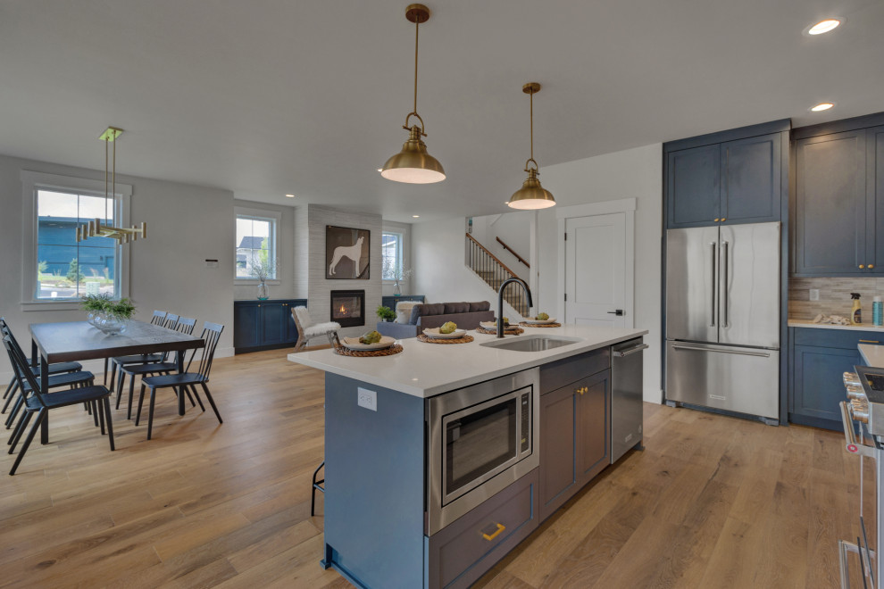 This is an example of a mid-sized modern l-shaped open plan kitchen in Portland with an undermount sink, recessed-panel cabinets, blue cabinets, quartz benchtops, white splashback, stone tile splashback, stainless steel appliances, light hardwood floors, with island and white benchtop.