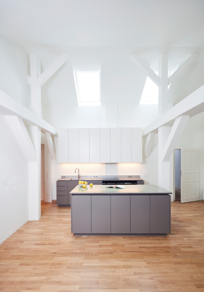 Expansive contemporary grey and white galley kitchen/diner in Munich with an integrated sink, flat-panel cabinets, grey cabinets, white splashback, black appliances, light hardwood flooring, an island, green worktops and exposed beams.