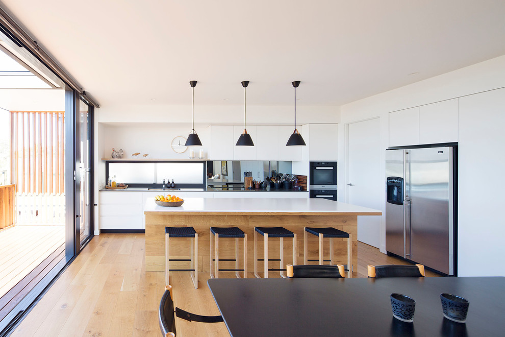 Large contemporary single-wall open plan kitchen in Sydney with flat-panel cabinets, white cabinets, quartz benchtops, metallic splashback, mirror splashback, stainless steel appliances, medium hardwood floors, with island and brown floor.