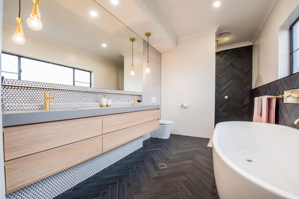 This is an example of a contemporary master bathroom in Sydney with engineered quartz benchtops, flat-panel cabinets, light wood cabinets, a freestanding tub, a one-piece toilet, white tile, mosaic tile, white walls, a wall-mount sink, black floor, an open shower and grey benchtops.