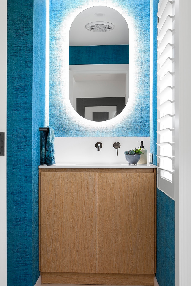 На фото: туалет среднего размера в современном стиле с плоскими фасадами, светлыми деревянными фасадами, унитазом-моноблоком, синей плиткой, керамической плиткой, бежевыми стенами, полом из керамической плитки, врезной раковиной, столешницей из искусственного кварца, бежевым полом, белой столешницей и встроенной тумбой