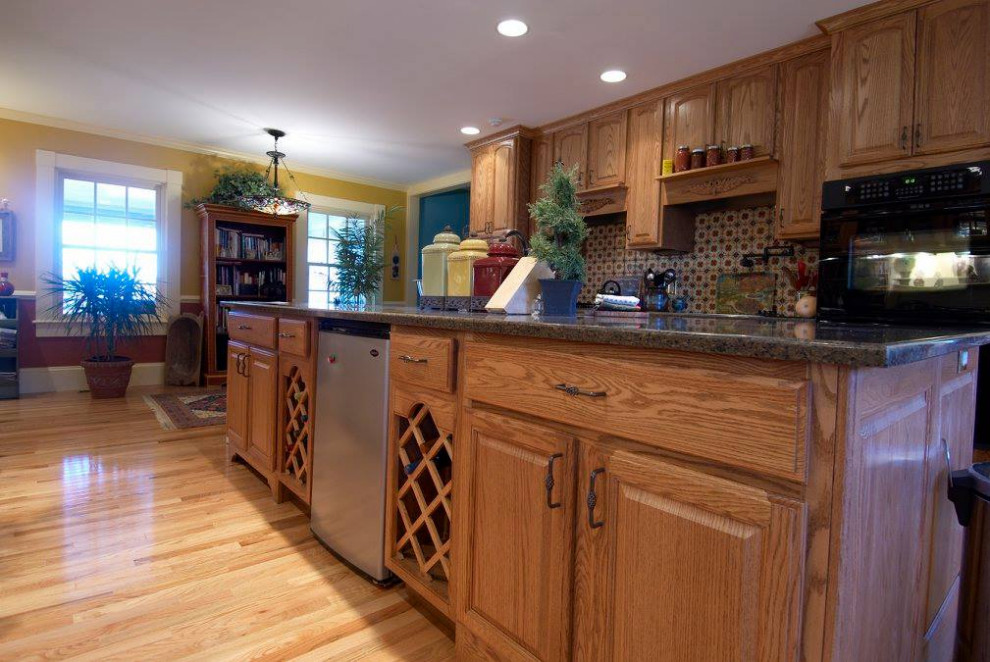 Example of a mid-sized single-wall light wood floor and brown floor kitchen design with recessed-panel cabinets, medium tone wood cabinets, brown backsplash, ceramic backsplash, stainless steel appliances, an island and gray countertops