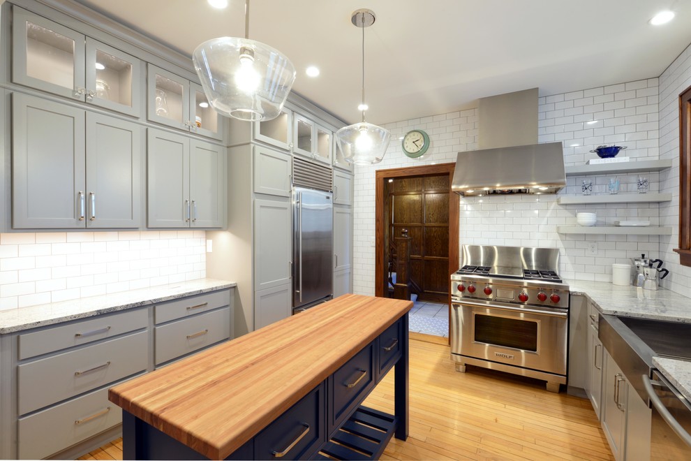 Mid-sized transitional light wood floor and brown floor enclosed kitchen photo with a farmhouse sink, shaker cabinets, gray cabinets, wood countertops, white backsplash, porcelain backsplash, stainless steel appliances, an island and brown countertops