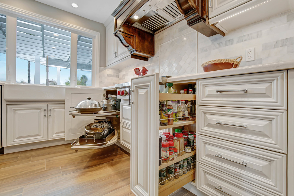 Photo of a mid-sized traditional l-shaped open plan kitchen in Los Angeles with a farmhouse sink, raised-panel cabinets, white cabinets, quartz benchtops, white splashback, marble splashback, stainless steel appliances, porcelain floors, with island, brown floor and white benchtop.