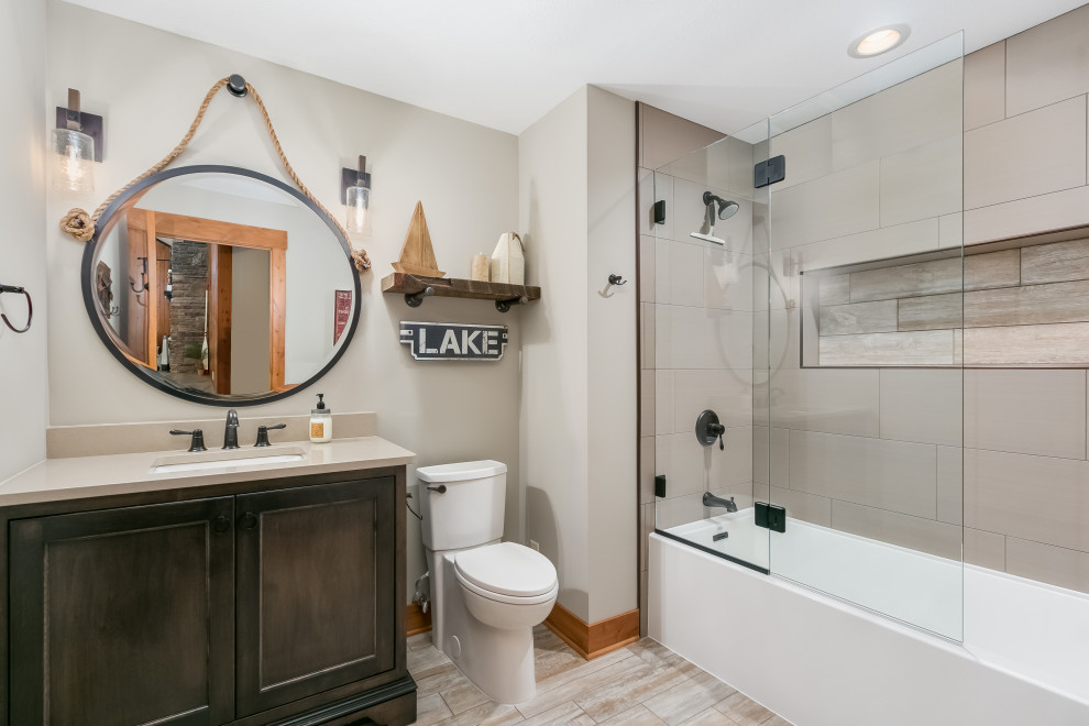 This is an example of a mid-sized beach style master bathroom in Minneapolis with flat-panel cabinets, dark wood cabinets, a drop-in tub, a shower/bathtub combo, a one-piece toilet, beige tile, ceramic tile, beige walls, laminate floors, a drop-in sink, marble benchtops, beige floor, a hinged shower door, multi-coloured benchtops, a double vanity and a floating vanity.
