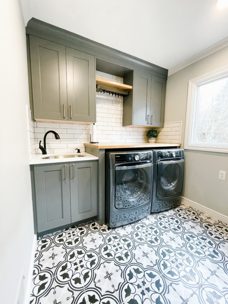 This is an example of a mid-sized country single-wall dedicated laundry room in Detroit with an undermount sink, shaker cabinets, grey cabinets, white splashback, subway tile splashback, grey walls, porcelain floors, a side-by-side washer and dryer, multi-coloured floor, wood benchtops and brown benchtop.