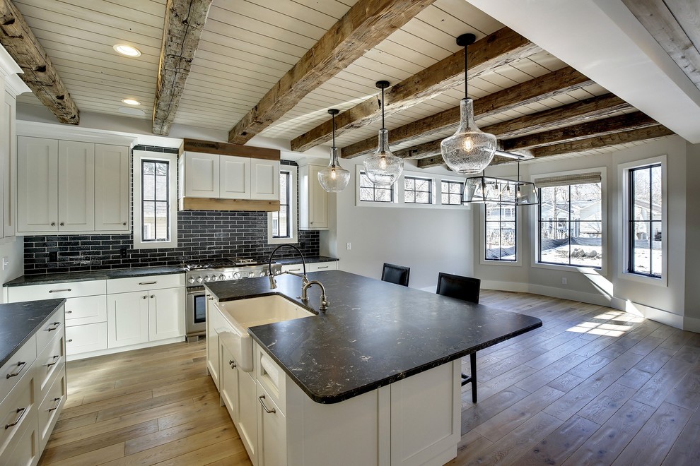 Mid-sized country l-shaped eat-in kitchen in Minneapolis with a farmhouse sink, shaker cabinets, white cabinets, quartzite benchtops, black splashback, subway tile splashback, panelled appliances, light hardwood floors and with island.