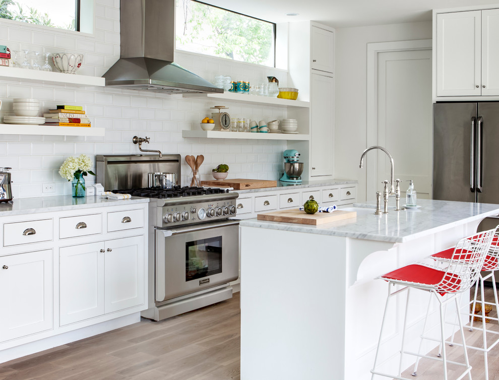 Inspiration for a transitional l-shaped kitchen in Austin with a farmhouse sink, beaded inset cabinets, white cabinets, white splashback, subway tile splashback, stainless steel appliances, light hardwood floors and with island.