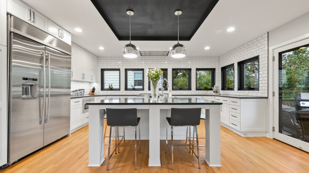 This is an example of a transitional u-shaped kitchen in Kansas City with shaker cabinets, white splashback, subway tile splashback, stainless steel appliances, light hardwood floors, with island and black benchtop.