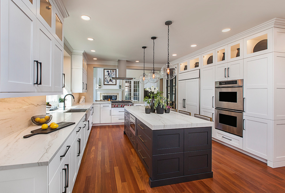 Large transitional l-shaped eat-in kitchen in Detroit with a farmhouse sink, shaker cabinets, white cabinets, quartzite benchtops, white splashback, stainless steel appliances, medium hardwood floors, with island, brown floor and white benchtop.