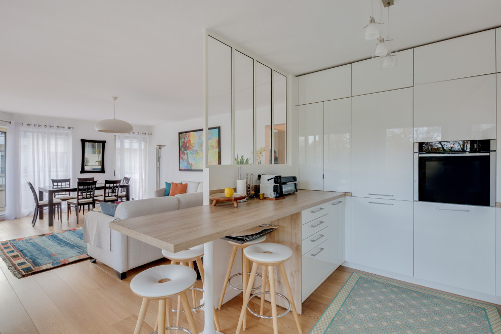 Photo of a scandinavian l-shaped kitchen in Paris with a single-bowl sink, beaded inset cabinets, white cabinets, wood benchtops, white splashback, porcelain splashback, black appliances, light hardwood floors, with island, blue floor and brown benchtop.