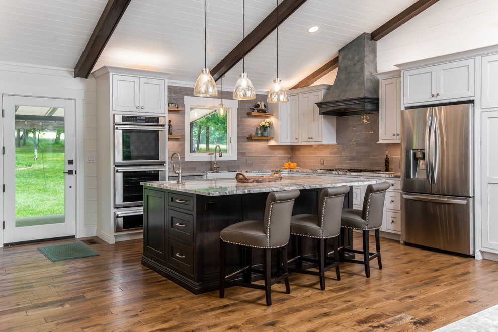 This is an example of a mid-sized country l-shaped open plan kitchen in Other with a farmhouse sink, recessed-panel cabinets, white cabinets, marble benchtops, grey splashback, subway tile splashback, stainless steel appliances, medium hardwood floors, with island, brown floor and white benchtop.
