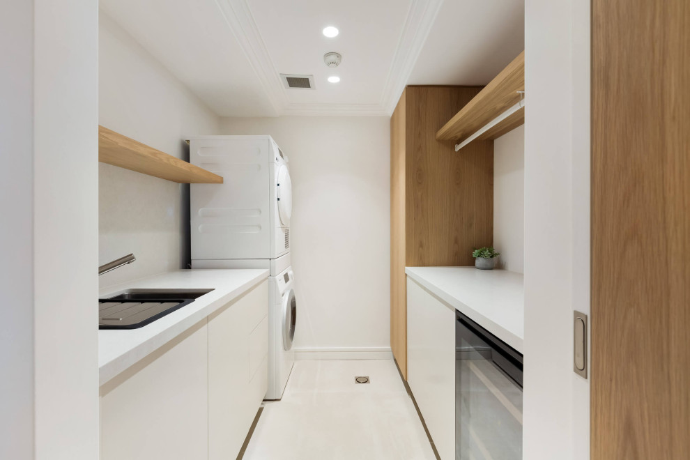 Photo of a mid-sized contemporary galley dedicated laundry room in Sydney with an undermount sink, light wood cabinets, marble benchtops, white splashback, marble splashback, white walls, ceramic floors, a stacked washer and dryer, beige floor and white benchtop.