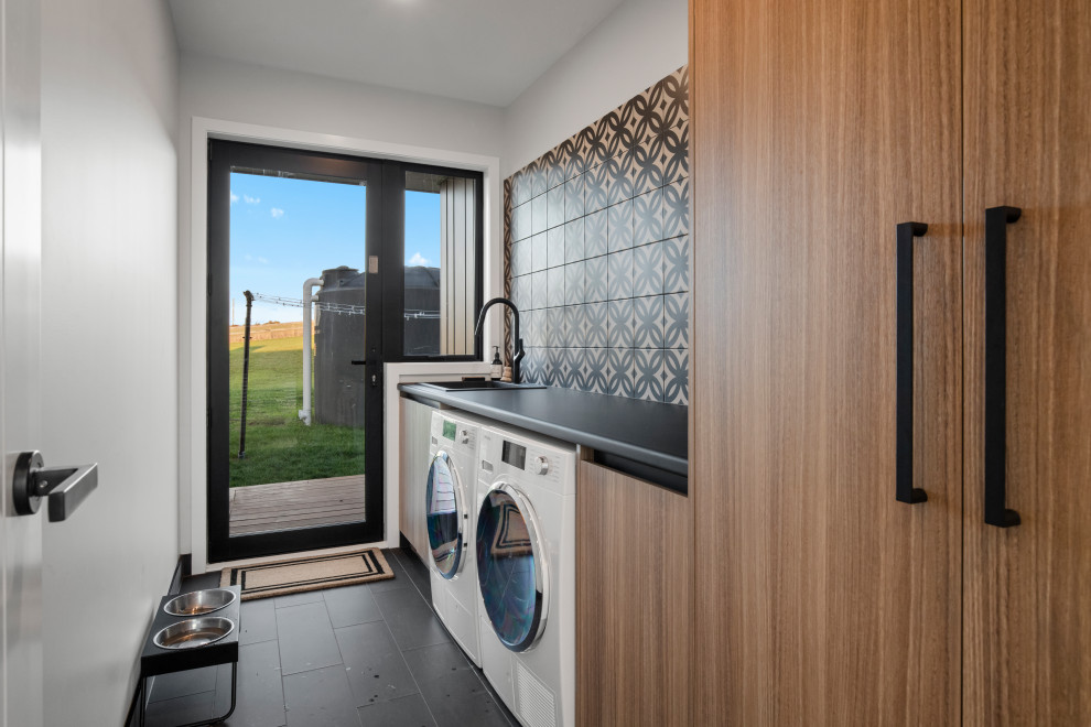 Photo of a modern dedicated laundry room in Auckland with laminate benchtops, multi-coloured splashback, mosaic tile splashback, porcelain floors, a side-by-side washer and dryer and black floor.