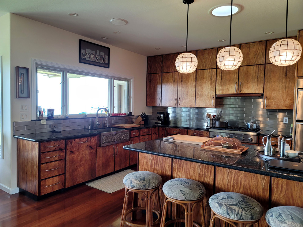 Photo of a modern eat-in kitchen in Hawaii with a farmhouse sink, flat-panel cabinets, light wood cabinets, granite benchtops, metallic splashback, metal splashback, stainless steel appliances, medium hardwood floors, with island and black benchtop.