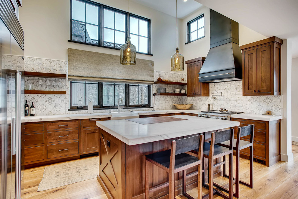 This is an example of a country l-shaped kitchen in Denver with a farmhouse sink, shaker cabinets, medium wood cabinets, multi-coloured splashback, stainless steel appliances, light hardwood floors, with island and white benchtop.