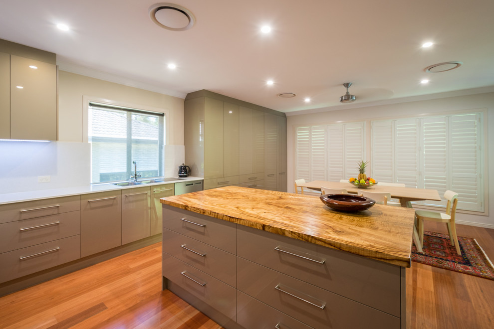 This is an example of an expansive contemporary l-shaped open plan kitchen in Gold Coast - Tweed with a drop-in sink, flat-panel cabinets, quartz benchtops, white splashback, stone slab splashback, stainless steel appliances and with island.