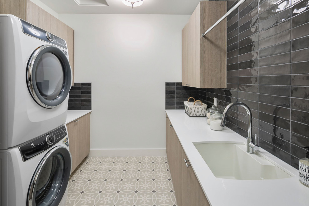 This is an example of a large galley dedicated laundry room in Seattle with an utility sink, flat-panel cabinets, grey cabinets, quartz benchtops, black splashback, subway tile splashback, white walls, ceramic floors, a stacked washer and dryer, multi-coloured floor and white benchtop.