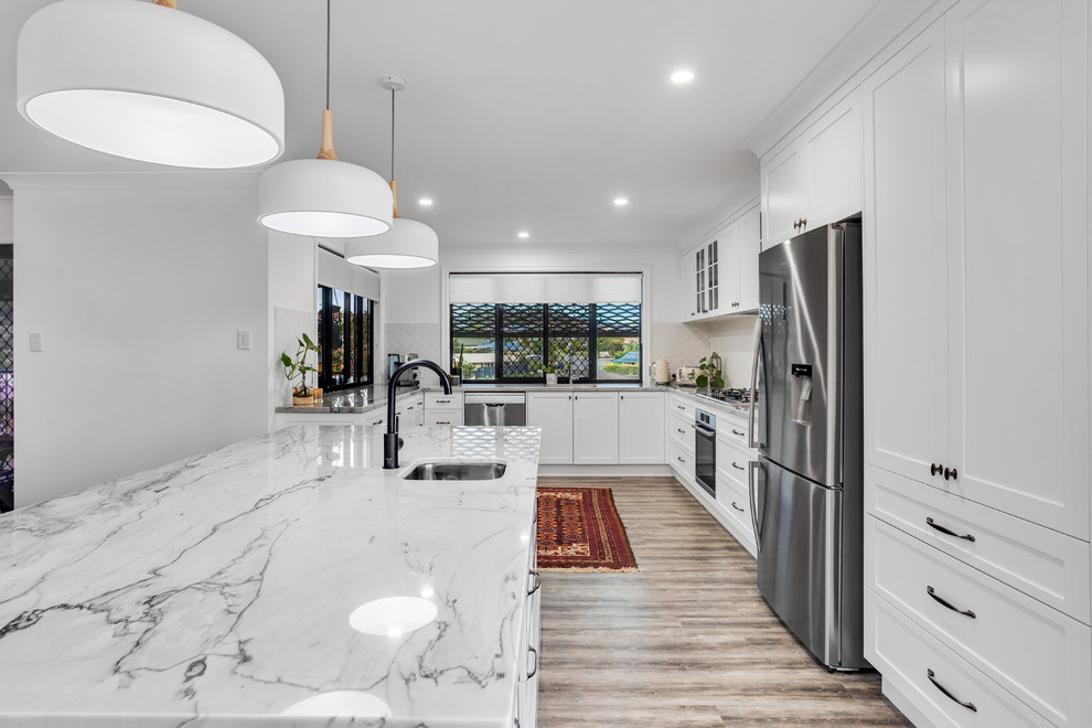 Large transitional u-shaped eat-in kitchen in Sunshine Coast with an undermount sink, shaker cabinets, white cabinets, quartzite benchtops, white splashback, ceramic splashback, stainless steel appliances, vinyl floors, with island and white benchtop.