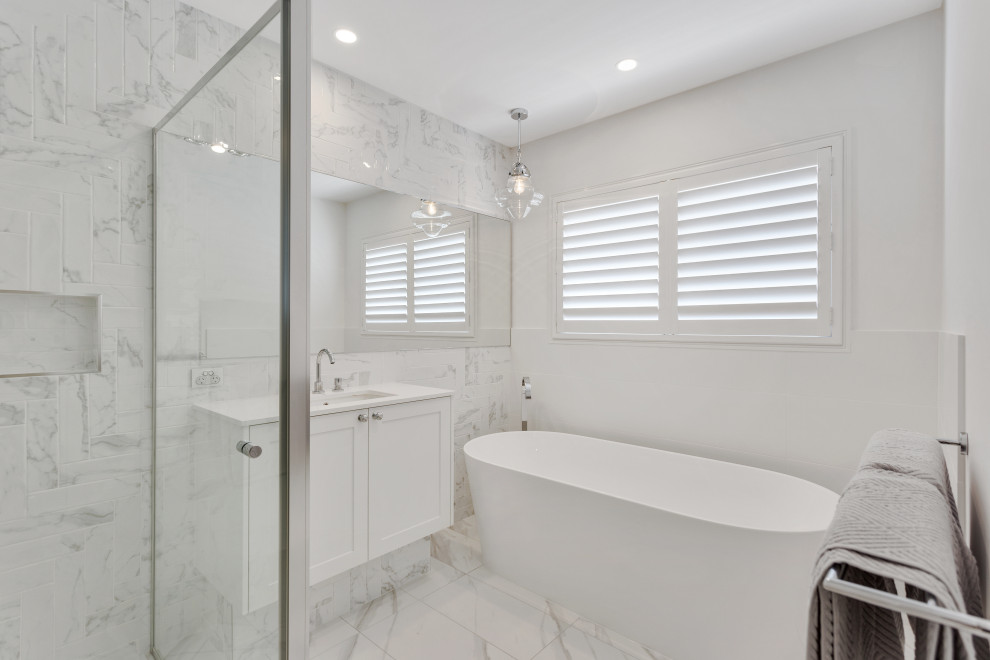 Photo of a large beach style 3/4 bathroom in Brisbane with shaker cabinets, white cabinets, a freestanding tub, multi-coloured tile, ceramic tile, white walls, ceramic floors, an undermount sink, engineered quartz benchtops, white floor, a hinged shower door, white benchtops, a niche, a single vanity and a floating vanity.