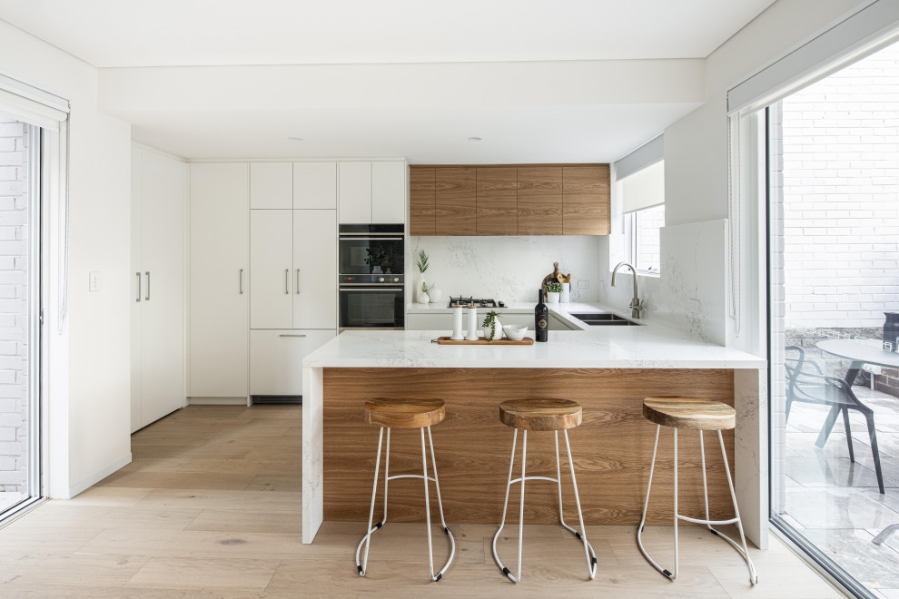 Foto på ett mellanstort funkis vit kök, med en dubbel diskho, släta luckor, vita skåp, bänkskiva i kvarts, vitt stänkskydd, svarta vitvaror, laminatgolv, en halv köksö och brunt golv